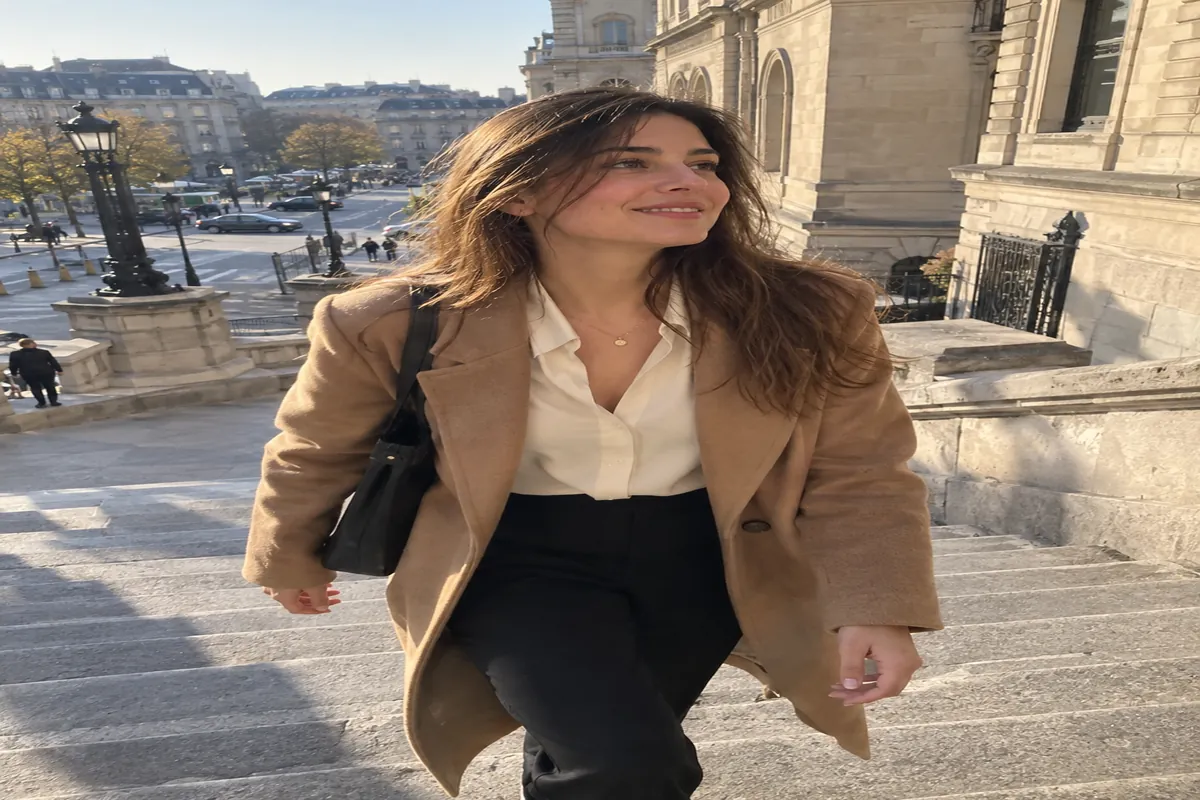 A woman in a camel coat and cream blouse walks up a stone staircase in morning light