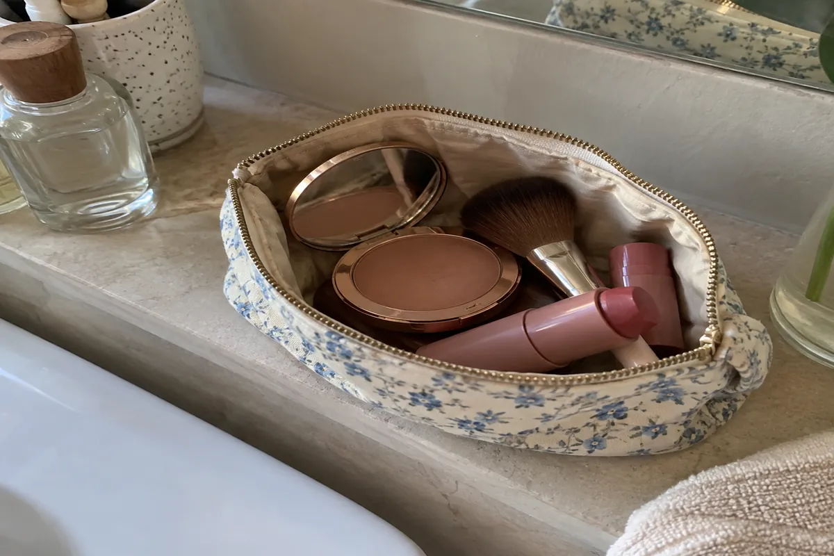 A small makeup bag open on a bathroom shelf with a compact, a brush and a tinted balm