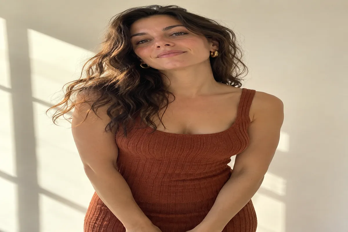 A woman in a rust knit dress and gold earrings stands in a sunlit studio, looking at the camera with a calm expression