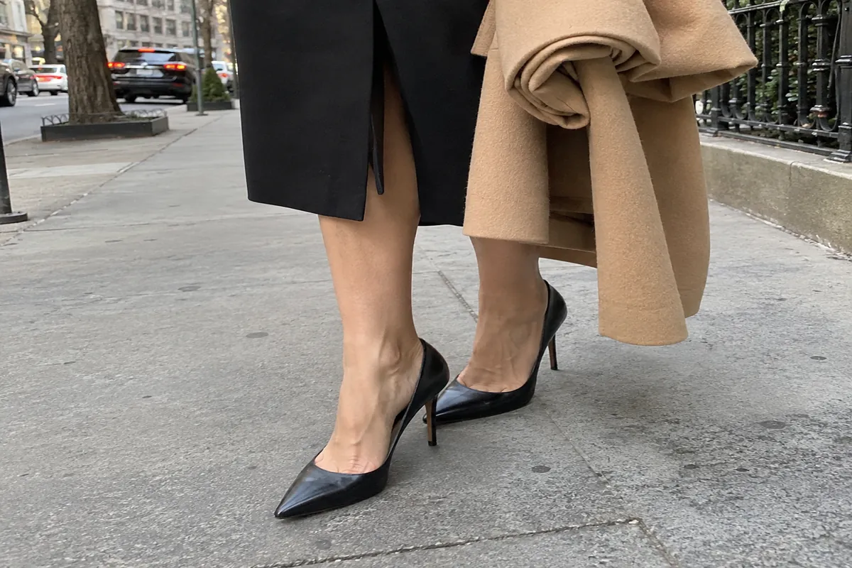 A woman's feet in black pumps standing on a city sidewalk, with a rolled-up coat under one arm